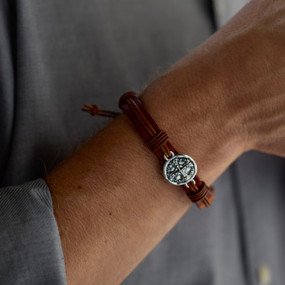 Close up of a man wearing a brown leather bracelet with a silver round charm with a cross on it.