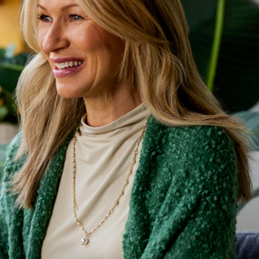 A smiling woman wearing a green wrap and a gold chain long necklace with a large white bead.