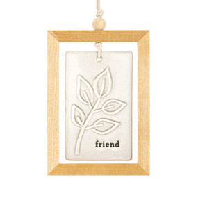 A hanging cream ceramic plaque with a leafy branch and the word "friend" suspended inside a light wood open frame.