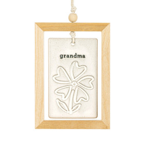 A hanging cream ceramic plaque with a flower and the word "grandma" suspended inside a light wood open frame.
