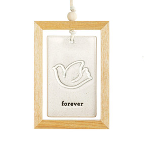 A hanging cream ceramic plaque with a dove and the word "forever" suspended inside a light wood open frame.