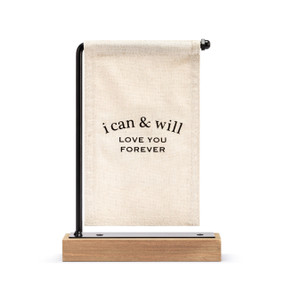 A cream canvas sign with black writing that says "i can & will Love You Forever" hanging on a black metal stand with a wood base.