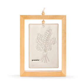 A cream ceramic plaque with a bouquet of flowers and the word "grateful" suspended inside a light wood open frame.
