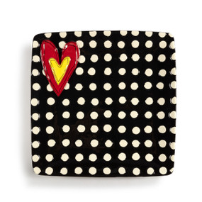 A square black ceramic plate with white polka dots and a raised red and yellow heart in the upper left corner.