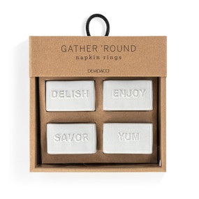 A set of four white ceramic square napkin rings. Each ring has a different word on it "Delish, Enjoy, Savor and Yum", displayed in a packaging box.