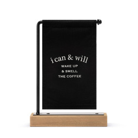A black canvas sign with white writing that says "i can & will Wake Up & Smell The Coffee" hanging on a black metal stand with a wood base.
