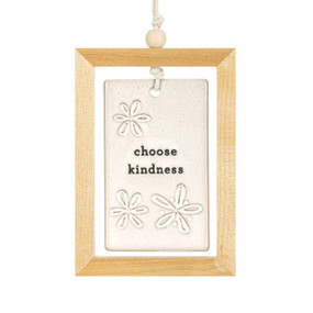 A hanging cream ceramic plaque with flowers and the saying "choose kindness" suspended inside a light wood open frame.