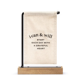 A cream canvas sign with black writing that says "i can & will Start Each Day With A Grateful Heart" hanging on a black metal stand with a wood base.