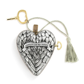 A silver heart shaped musical sculpture with a wing pattern that reads "always in my heart". The heart has a silver tassel and gold key attached.