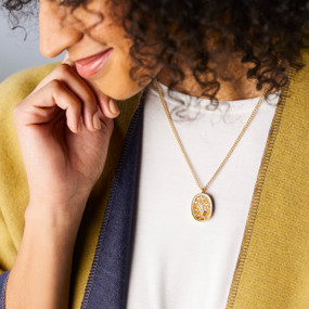 A woman wearing a gold chain necklace with an oval charm that has mustard seeds and a silver tree inside.