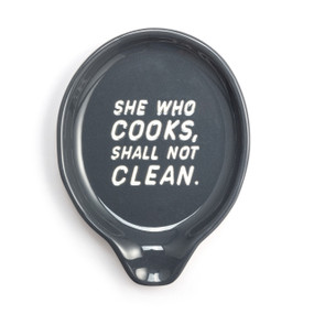 Top down view of a gray ceramic spoon rest that says "She Who Cooks, Shall Not Clean" in white.
