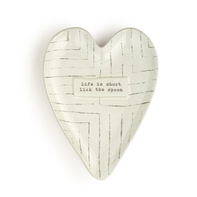 An off white heart shaped ceramic spoon rest with thin lines and the saying "life is short lick the spoon" on it.