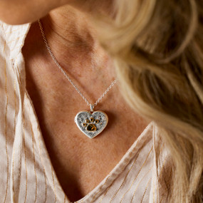 Close up of a woman wearing a silver chain necklace with a heart shaped charm with a gold paw print on it.