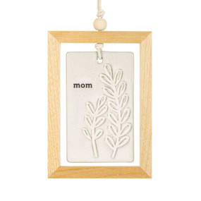 A hanging cream ceramic plaque with leaves and the word "mom" suspended inside a light wood open frame.