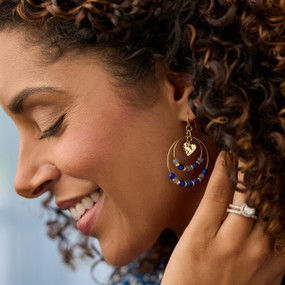 Close up of a woman wearing dual hoop earrings with blue and gray beads and a gold heart charm in the middle.