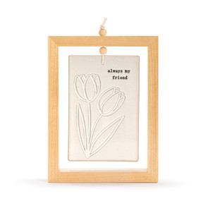 A cream ceramic plaque with tulips and the saying "always my friend" suspended inside a light wood open frame.
