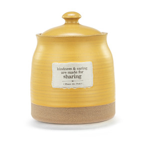 A yellow round canister with a lid and textured tan base. There is a patch on the front that says "kindness & caring are made for sharing".