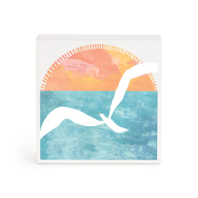 A small white box artwork of a white bird in flight over a blue ocean and setting sun.