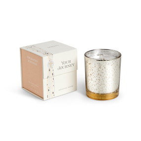 A candle in a white and gold round glass container sitting next to a peach and white packaging box. The candle name is Enchanted Mornings.