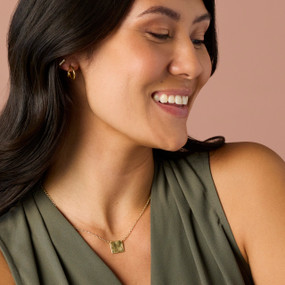 A smiling woman wearing a gold chain necklace with a square charm of a daisy flower.