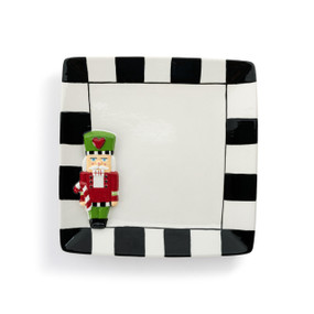 A square ceramic plate with a black and white check border and a raised red and green nutcracker on the bottom left corner.