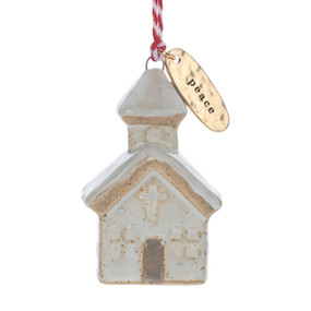 A small ceramic cream church shaped ornament with snow on the roof and a gold metal tag at the top with the word "peace".