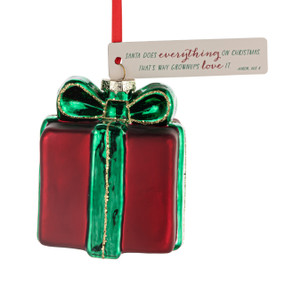 A blown glass ornament of a red present wrapped in a green bow. There is a cream tag with the saying "Santa does everything on Christmas that's why grownups love it".