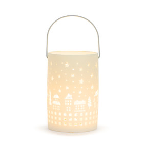 A white round ceramic lantern with a rope handle and a cut out pattern of a holiday village displayed lit.