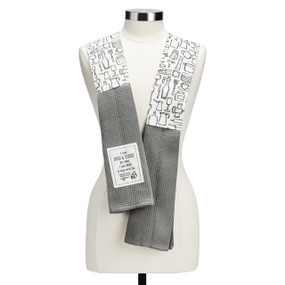 A white fabric kitchen boa with kitchen tool illustrations and gray towel ends. There is a fabric patch that says "I love hugs and kisses but what I love most is help with the dishes".