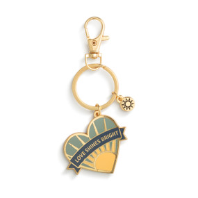 A gold keychain with two charms. One is a small round sun and the other is shaped like a heart and has the saying "Love Shines Bright".