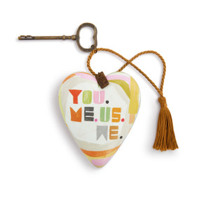 A white heart shaped sculpture with muted colors and the saying "You. Me. Us. We.". The heart has a metal key and gold tassel.