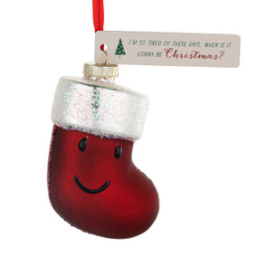 A blown glass ornament of a red stocking with a smiling face. There is a cream tag with the saying "I'm so tired of these days. When is it gonna be Christmas?".