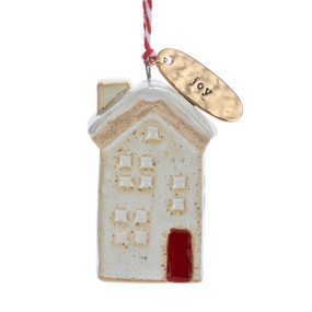 A small ceramic cream house shaped ornament with snow on the roof and a gold metal tag at the top with the word "Joy".