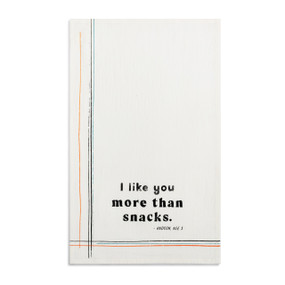 A white kitchen towel with the saying "I like you more than snacks" displayed laid out flat.