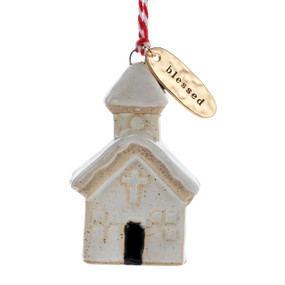 A small ceramic cream church shaped ornament with snow on the roof and a gold metal tag at the top with the word "blessed".