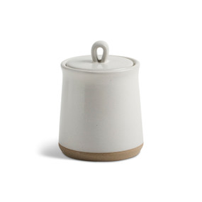 A himalayan citrus candle in a tall white ceramic jar with a lid and tan textured base.