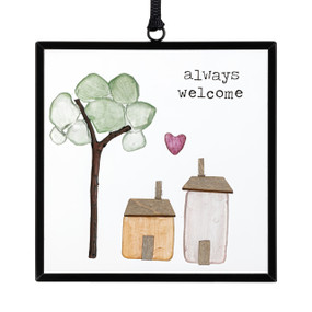 A small square suncatcher with two houses and a tree made out of small stones and the saying "always welcome".