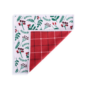 A double sided square dish cloth that is white with holly illustrations on one side and red plaid on the other displayed with the red corner turned back.