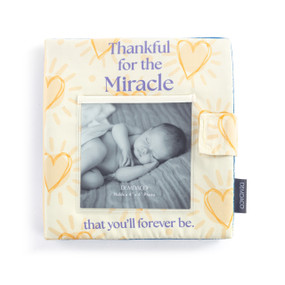 A light yellow fabric baby book with an opening on the front to hold a photo. The name of the book is "Thankful for the Miracle that you'll forever be."