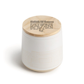 A white ceramic jar candle. The lid is wood with the saying "RE: Out of the Office Searching for sasquatch" with a short write up about someone hiking in the woods.