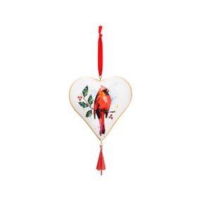 A heart shaped tan ornament with a watercolor painted image of a red cardinal sitting on a holly branch painted on the center. With a red ribbon at the top and bottom of the ornament.
