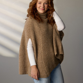 A woman wearing a taupe turtleneck pullover cape stands with her body angled to the right and her head turned to the front Her right arm is at her side, her left arm is bent with her hand behind her ear, in order to show the arm openings in the cape.