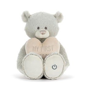 Front view of a grey teddy bear. The bear is holding a cream-colored heart in its lap, with the words "my first teddy" printed across the center. A power button symbol is on the left foot.