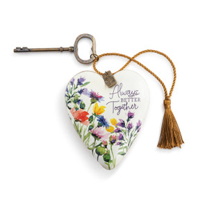 Top down view of a white and gold art heart. The front of the heart has a design of wildflowers on a white background. The phrase "Always Better Together" is printed in purple script on the heart's upper left side. A gold cord with a tassel at the end is attached to the top and displayed to the heart's left. The cord is threaded through a bronze key that can be attached at the back as a stand.