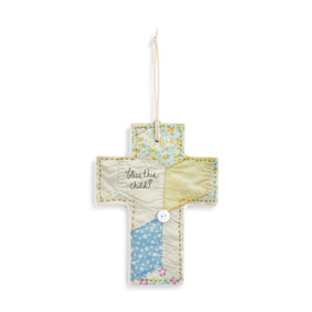 Top down view of a ceramic cross with a ribbon threaded through the top for hanging. The cross is decorated with patches of blue, green and natural color to resemble a patchwork quilt. The phrase "Bless This Child" is printed in typeface on the left side of the crossbar., with a small white button affixed on a corner of the patch near the center of the cross.
