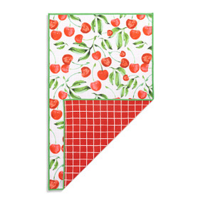 Top down view of a kitchen towel with a pattern of cherries on stems with green leaves on a white background on the front. The bottom right corner is folded up to show the back of the towel, which has a red and white plaid print.