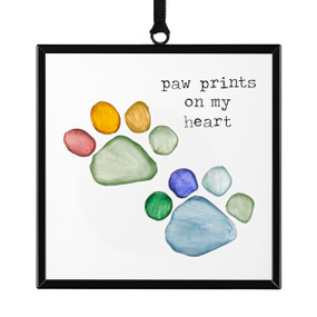 Front view of a clear glass suncatcher in a black frame, with a black ribbon threaded through a loop hanger on the top. On the surface of the glass is a set of animal pawprints in rainbow colors a set of animal pawprints in rainbow colors and the words "pawprints on my heart" in the upper right corner.