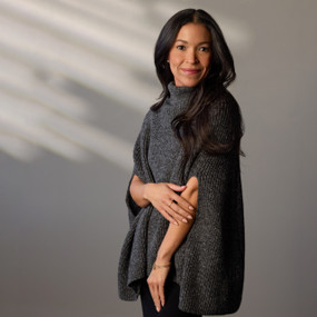 A woman wearing a black turtleneck pullover cape stands with her body turned to the left, her head turned to the front. Her left arm is at her side, her right arm folded over her waist.
