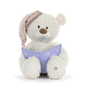 Front view of a cream-colored teddy bear. The bear is wearing a light brown night cap printed with stars and "zzz" over one ear.  The bear is also holding a purple fabric storybook. A power button symbol is on the left foot.