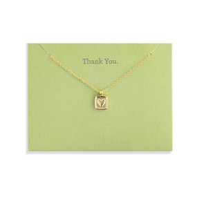 Top down view of a gold necklace and a square gold charm with a heart at its center attached to a green card. "Thank you" is printed at the top of the card above the necklace.